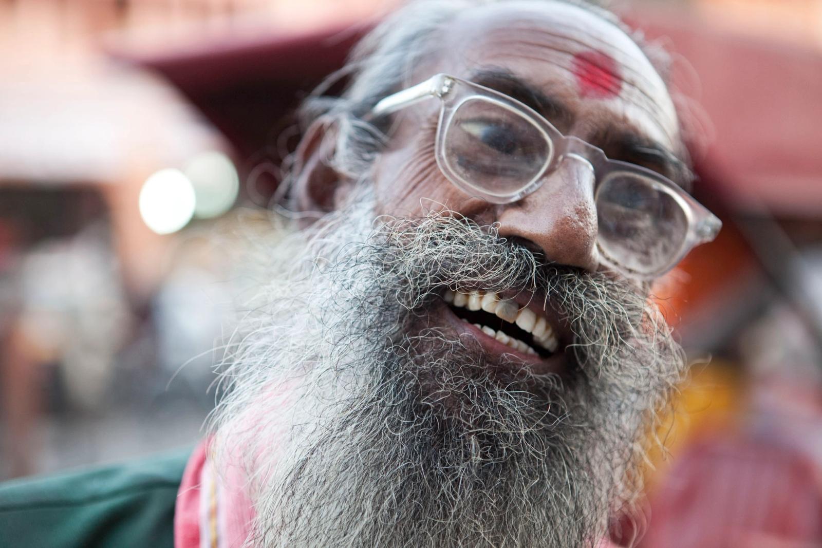A man with a long, grey beard and a red dot on his forehead, wearing a pink shirt and glasses, is smiling and looking directly at the camera.