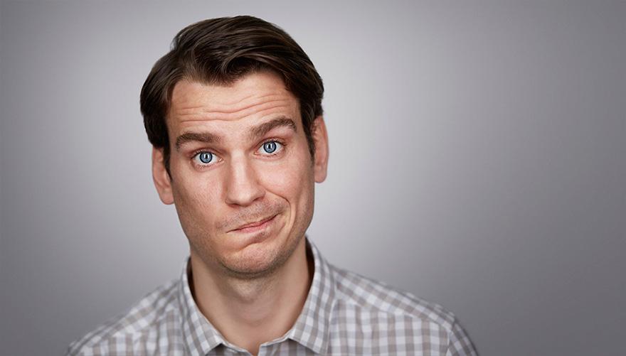 A man with a neutral expression, wearing a white and grey checkered shirt, looking directly at the camera with a slight smile.