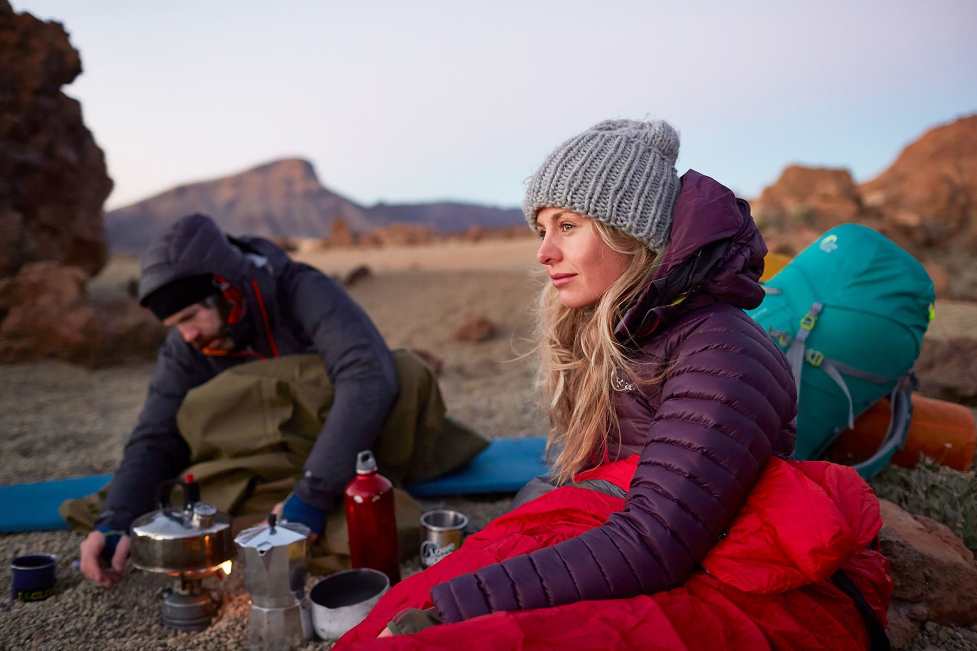 Two people, likely hikers or campers, sitting on a rocky outcrop, surrounded by camping gear and cooking a meal on a portable stove.