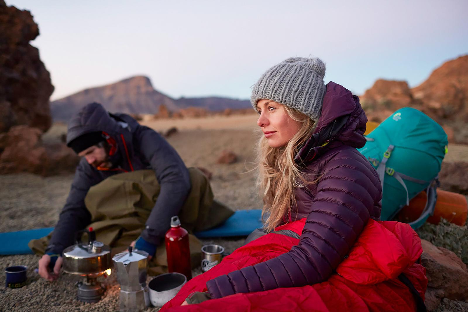 Two people, likely hikers or campers, sitting on a rocky outcrop, surrounded by camping gear and cooking a meal on a portable stove.
