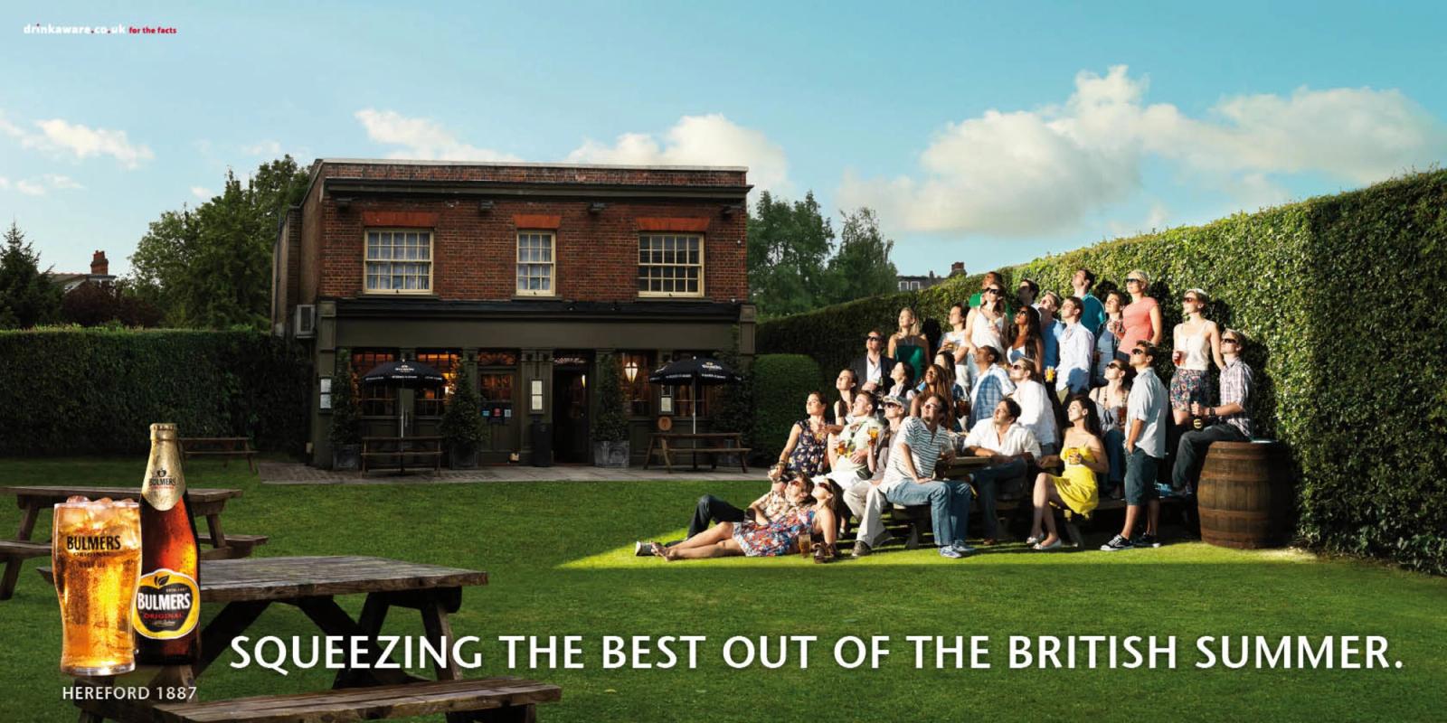 A group of people sitting on a grassy lawn in front of a pub, with a table and a beer bottle in the foreground.
