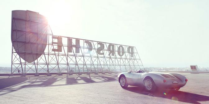 A 1955 Porsche 550 Spyder is parked in front of a large, metal, industrial-style sign reading 'Additive CGI' in a desert-like setting.