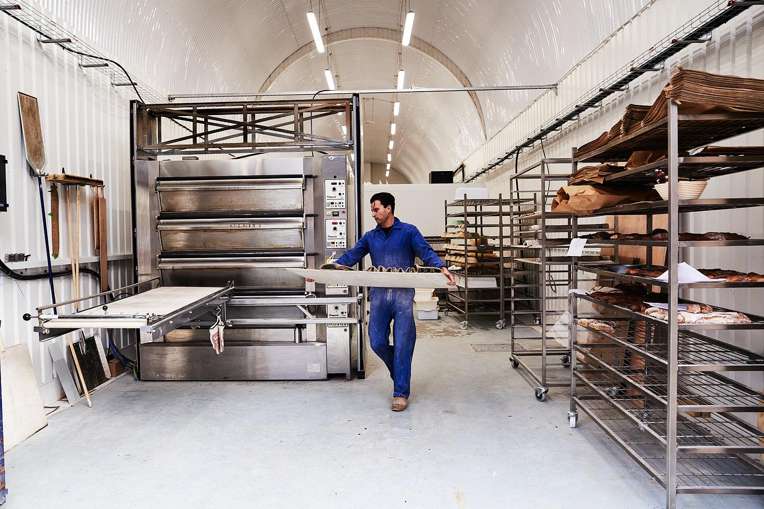 A man in a blue jumpsuit and a white apron is standing in a large industrial kitchen, surrounded by various food preparation equipment and ingredients.
