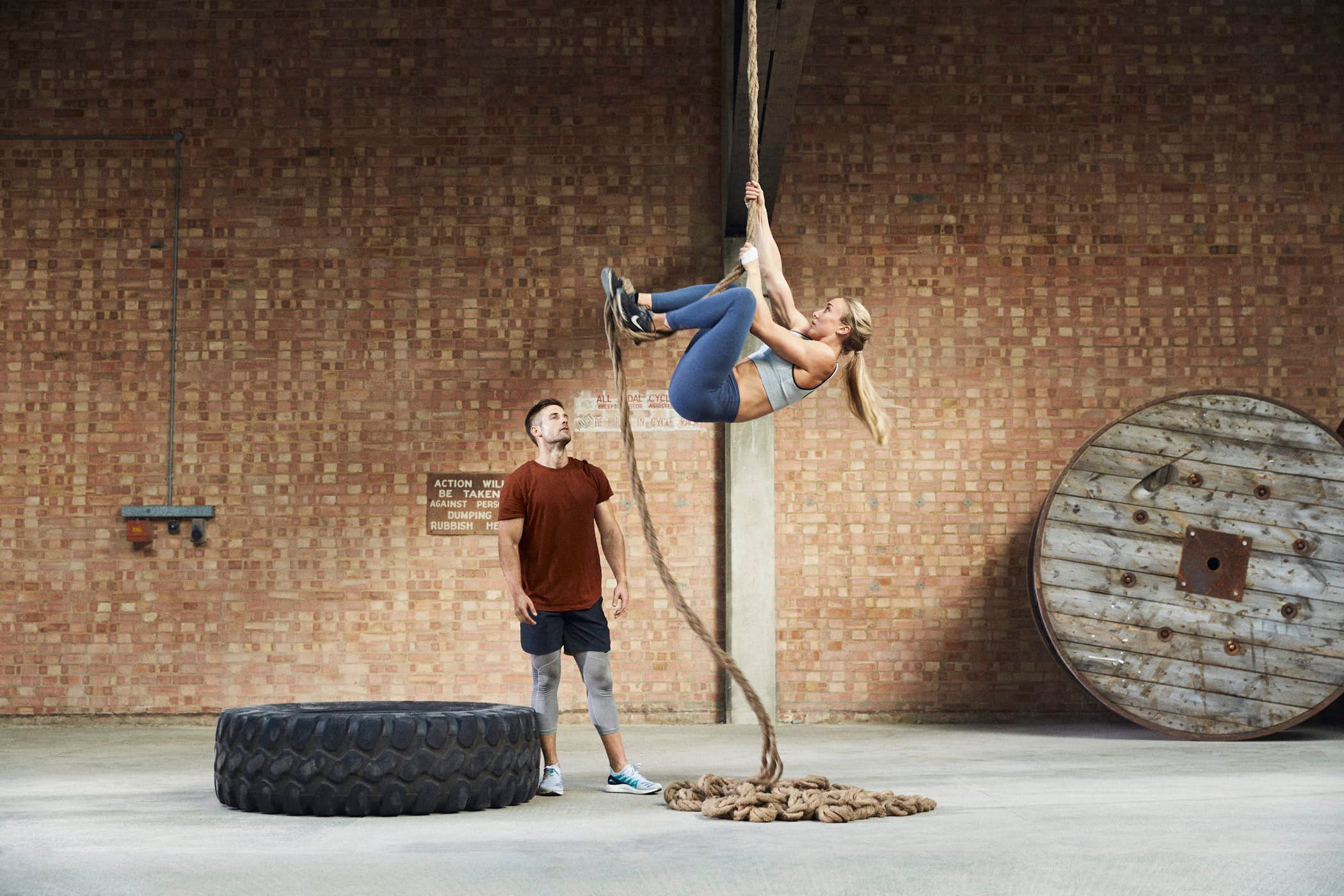A man and woman are in a gym, with the woman hanging from a rope. She is wearing a sports bra and blue leggings, and has her long blonde hair tied back. She is suspended in.