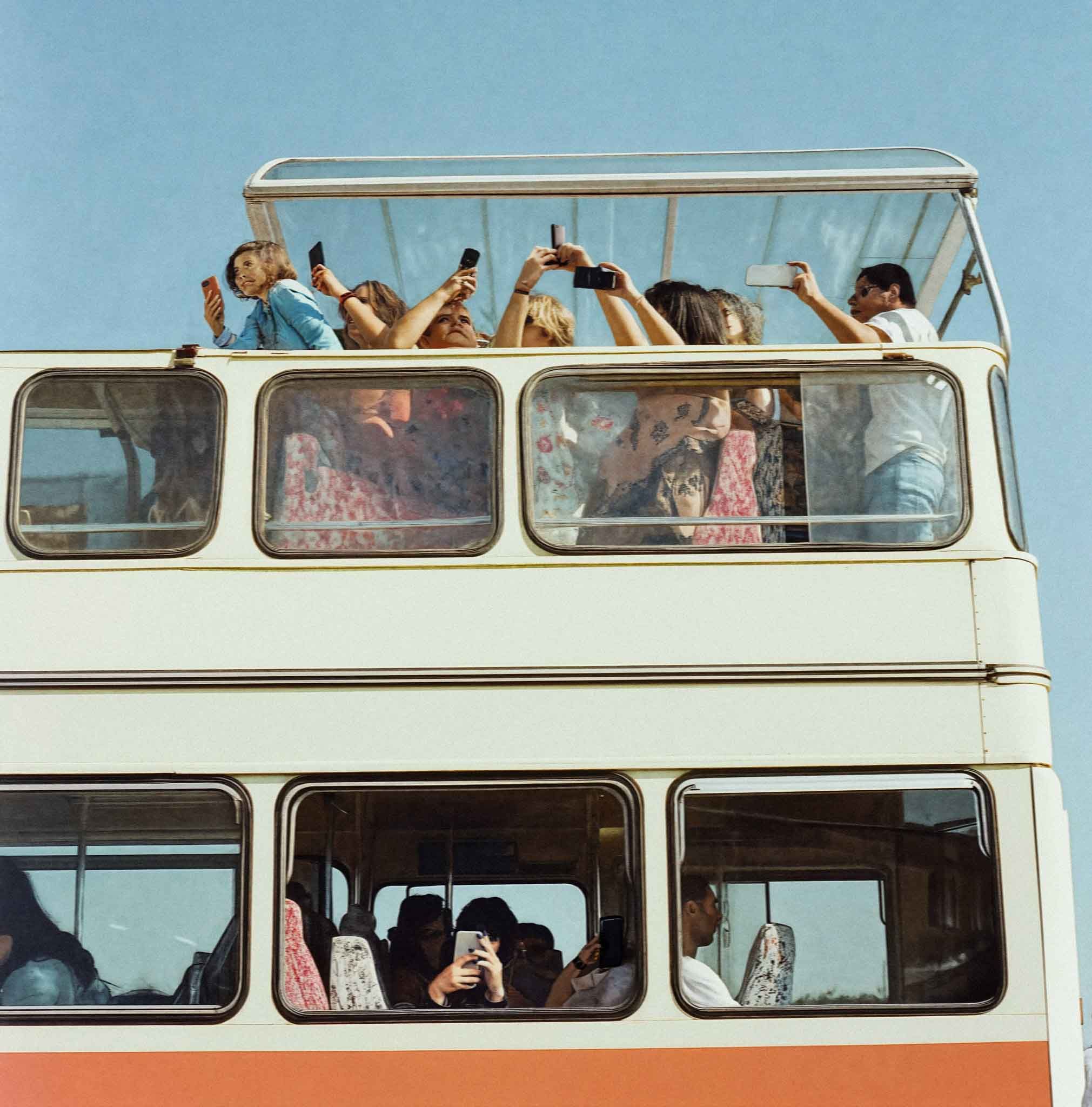 A crowd of people, mostly women, are standing on the upper deck of a double-decker bus, holding up their mobile phones to take photos of something outside the bus.