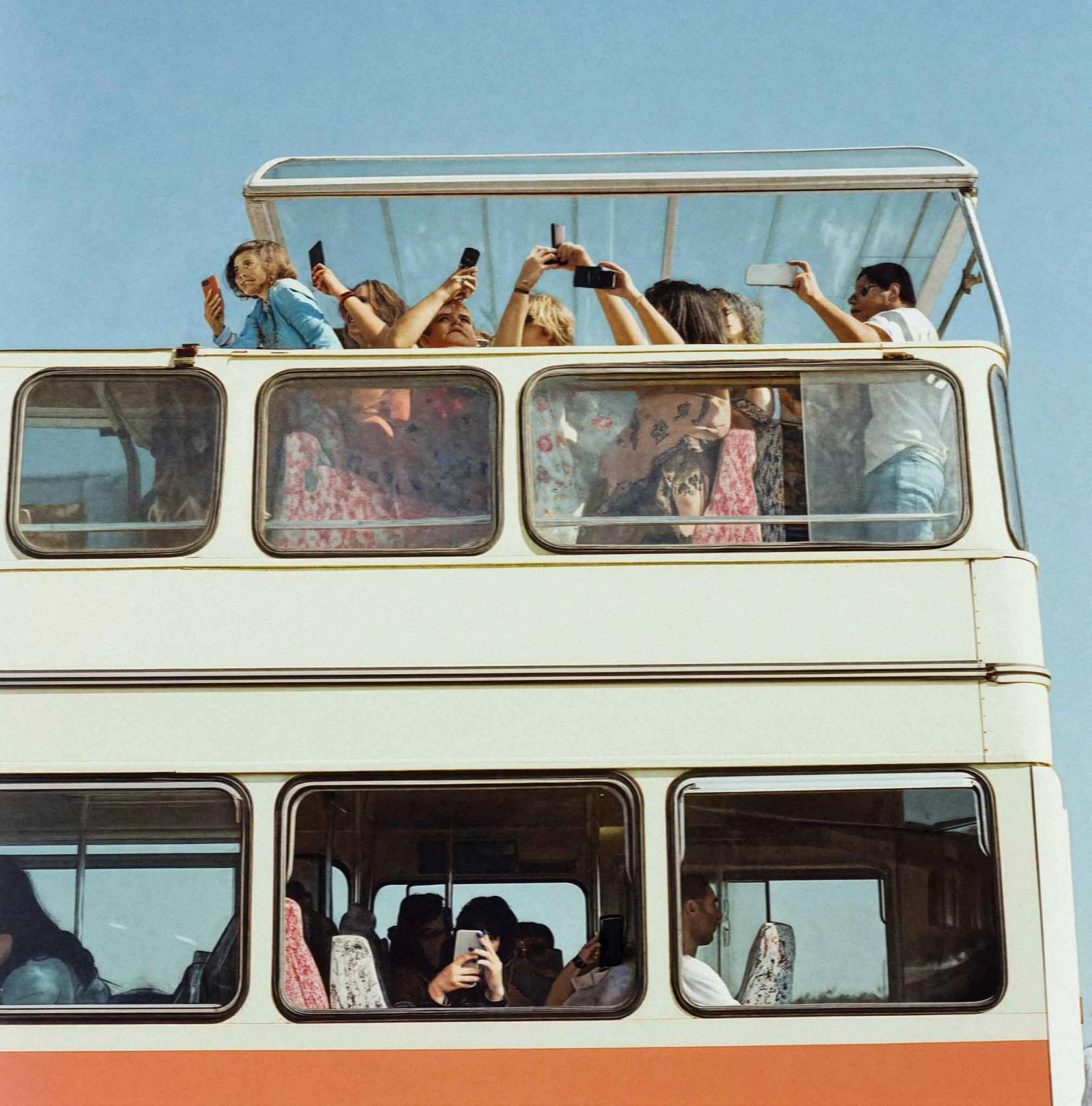A crowd of people, mostly women, are standing on the upper deck of a double-decker bus, holding up their mobile phones to take photos of something outside the bus.