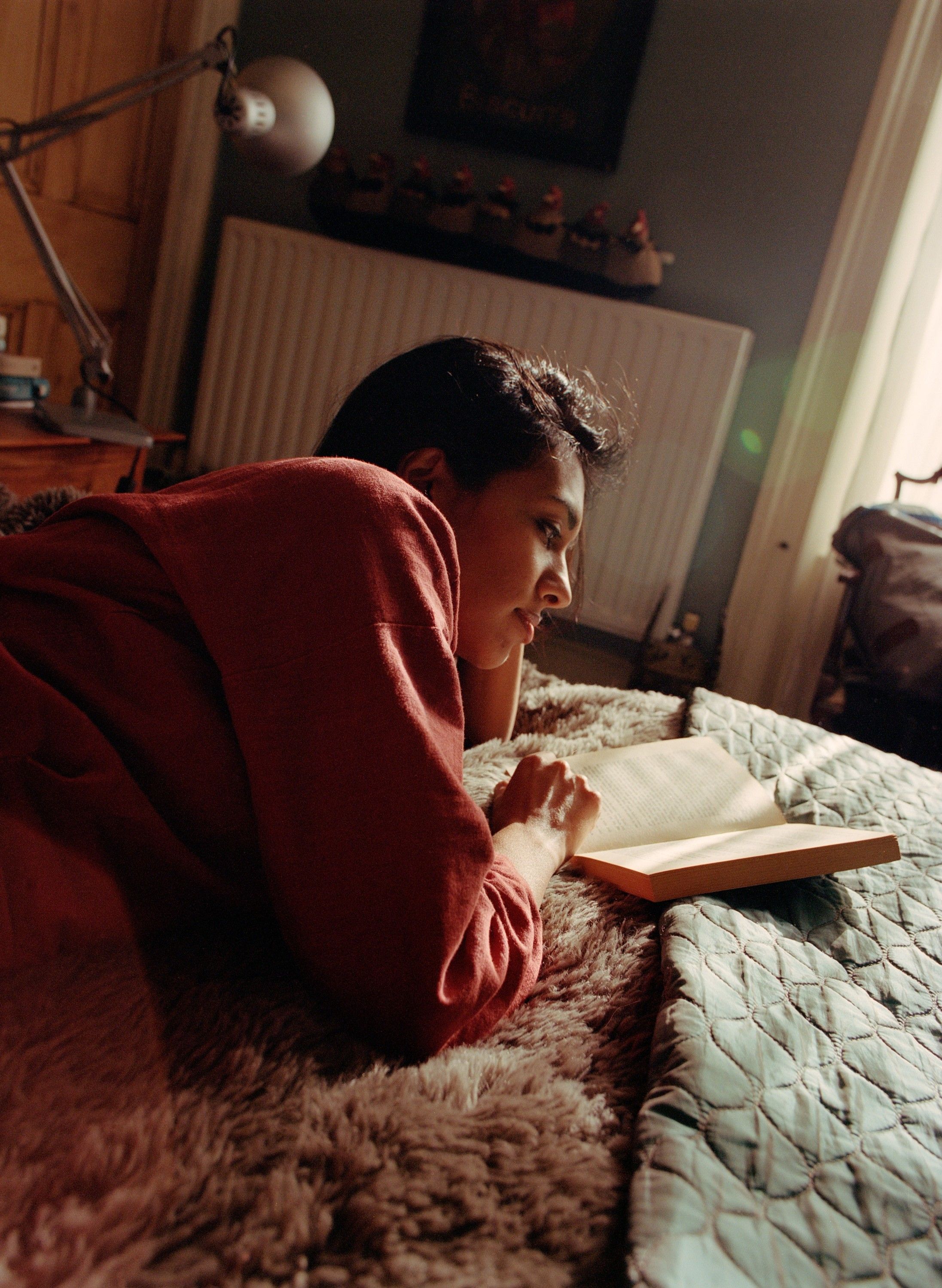 A woman in a red sweater lies on a bed, reading a book.