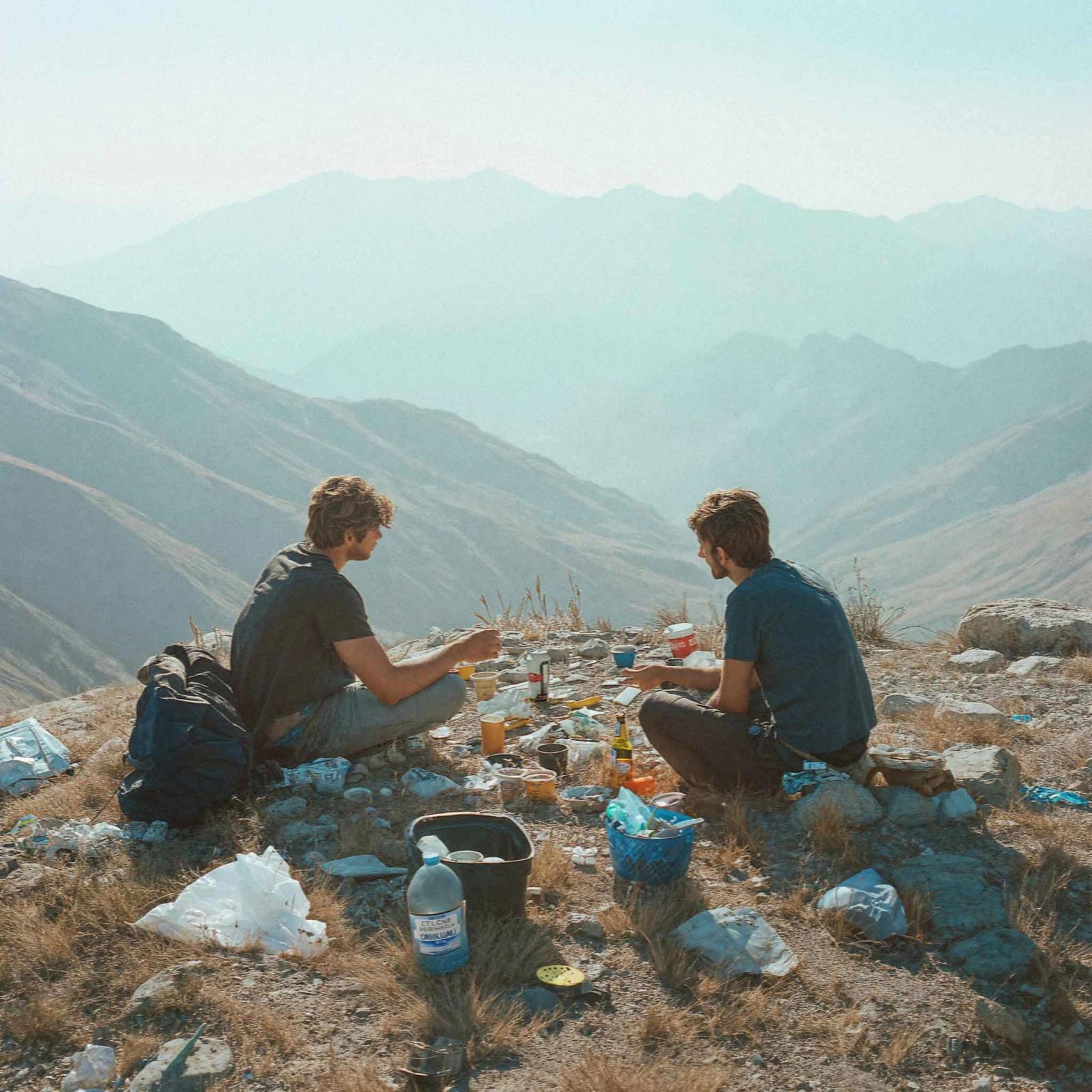 Two men sitting on a rocky outcrop, eating and drinking, with a mountainous landscape in the background.