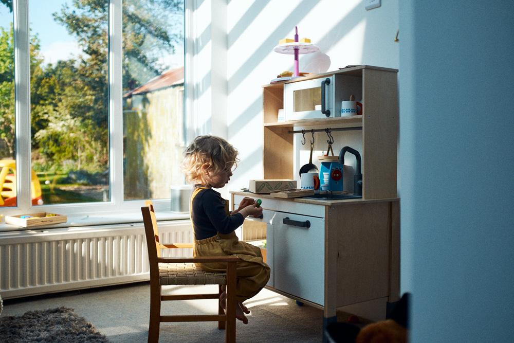 A young child sits in a small kitchen area, playing with a toy food item. The child is barefoot and wearing yellow overalls, and is facing a small kitchen area with a toy stove.