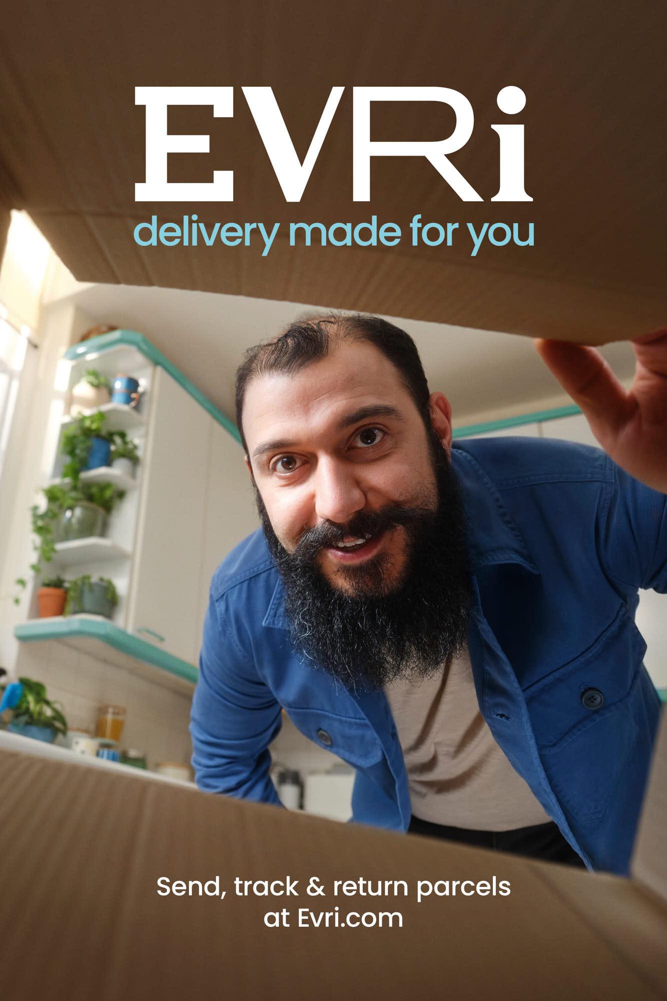 A man with a long black beard and a blue jacket peers into an open cardboard box, smiling at the camera. The box's contents are out of frame, but the man's expression suggests he.