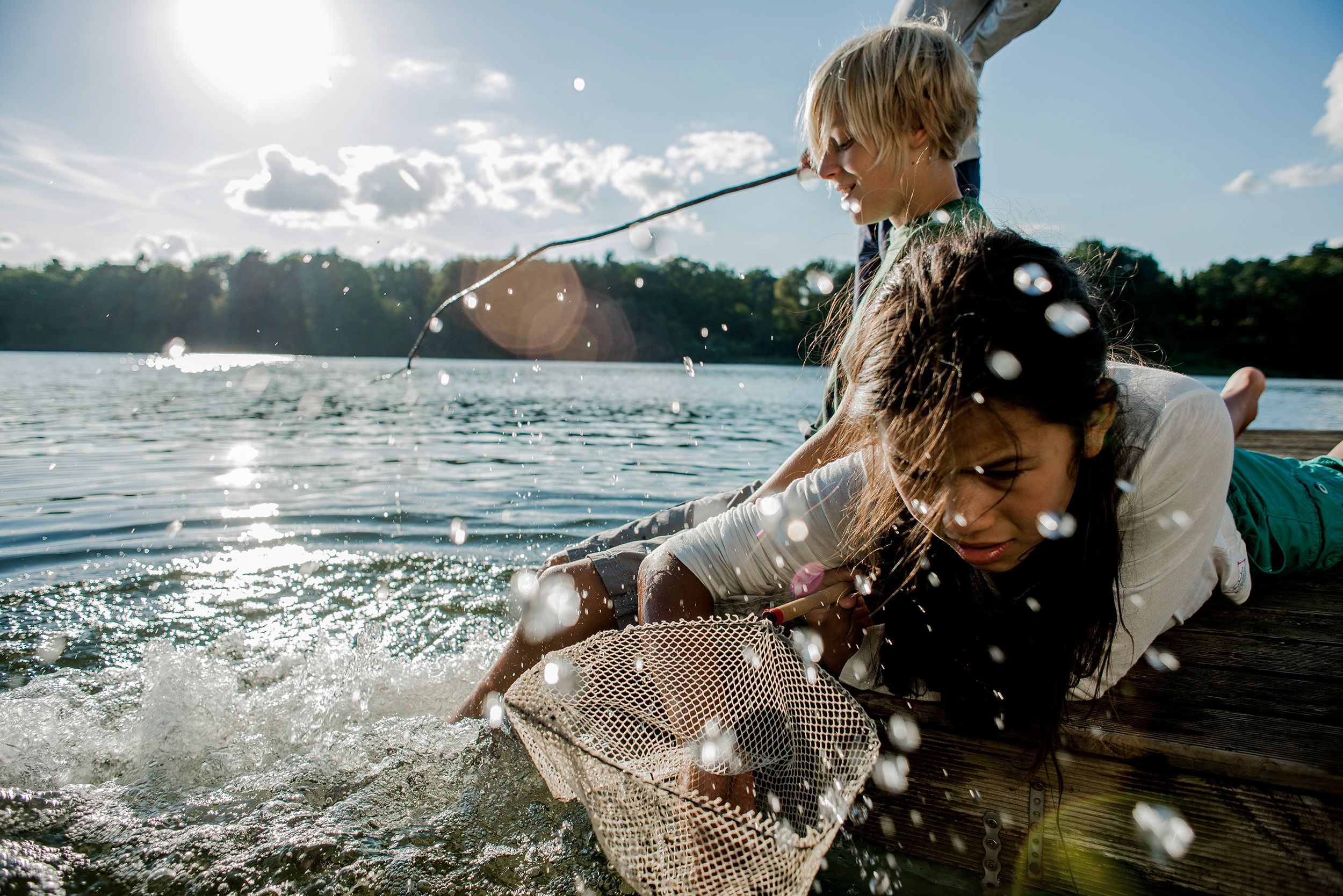 Two children, a boy and a girl, are fishing from a dock, with the boy holding a fishing rod and the girl holding a net, in a lake surrounded by trees and a sunny sky.