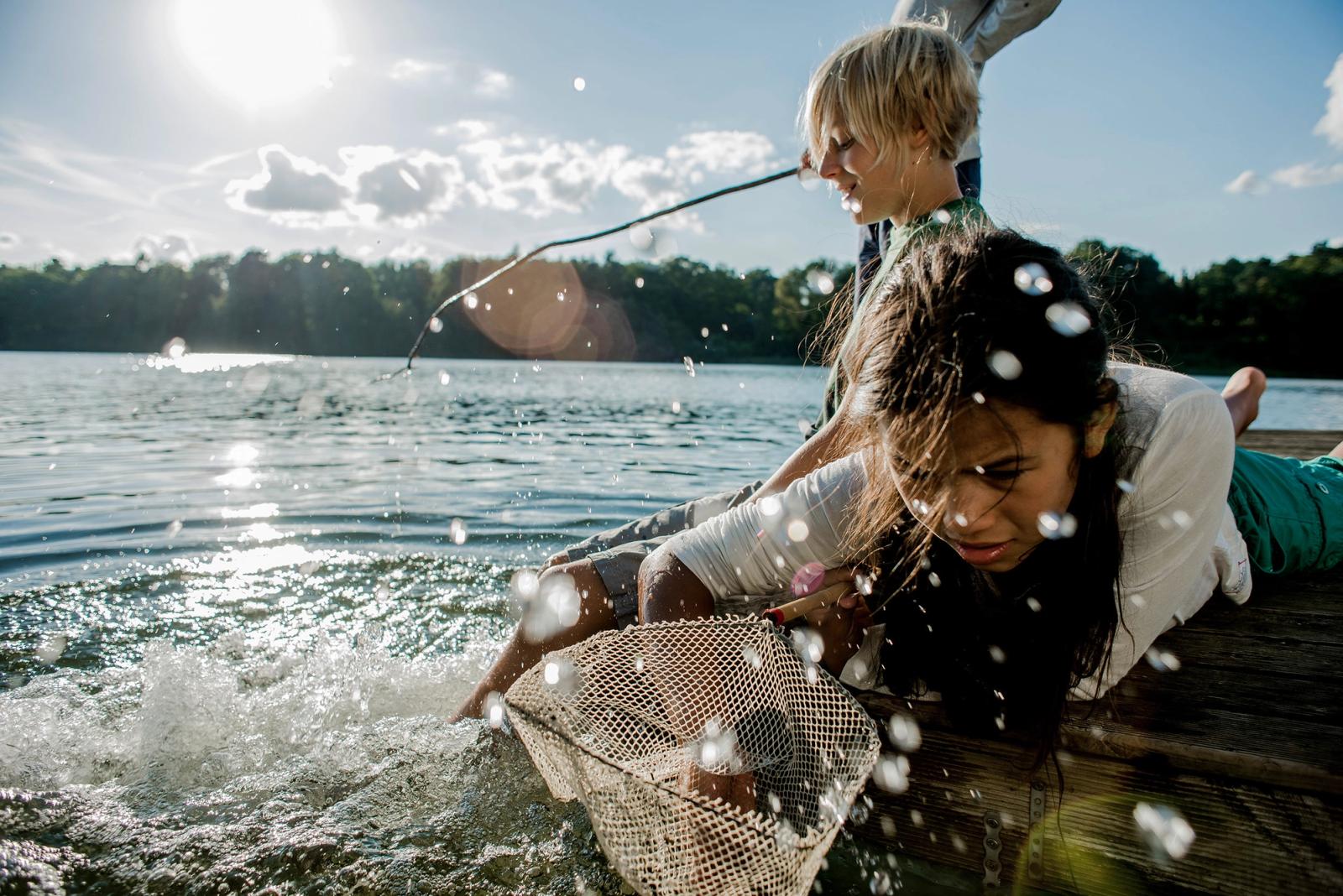 Two children, a boy and a girl, are fishing from a dock, with the boy holding a fishing rod and the girl holding a net, in a lake surrounded by trees and a sunny sky.