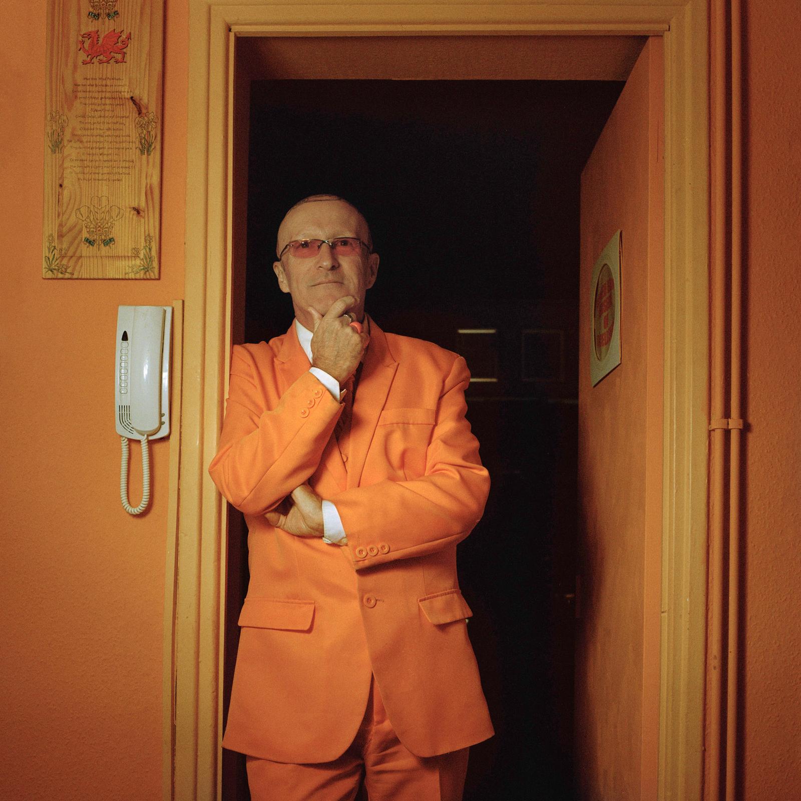 A man in a bright orange suit stands in a doorway, gazing into the distance.