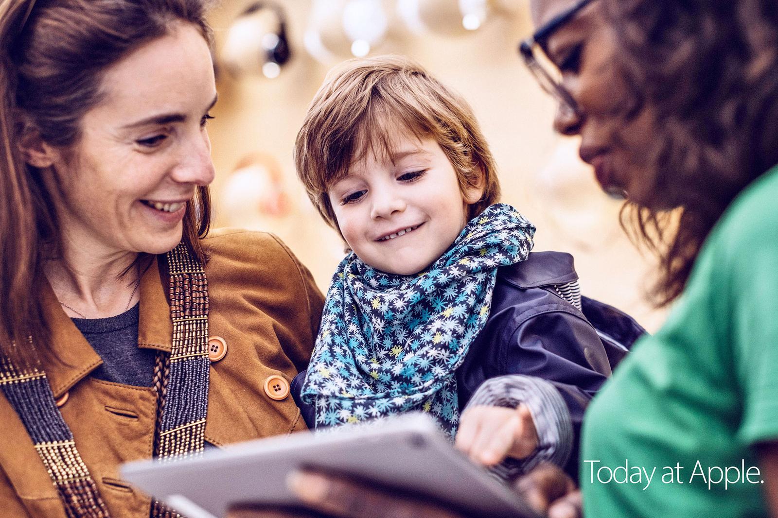 A woman, a child, and a woman in a green shirt are gathered around a tablet, with the child pointing at the screen.