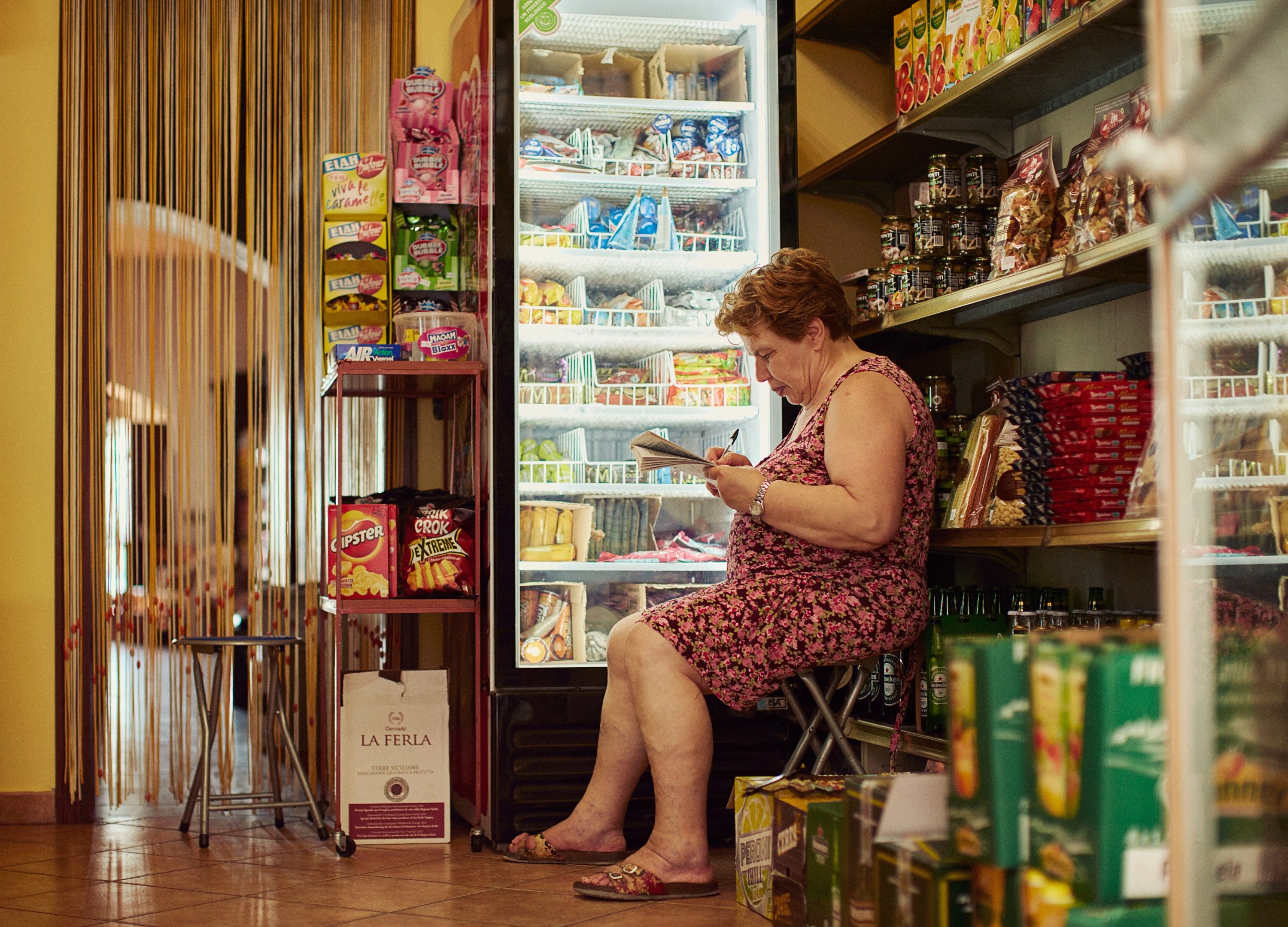 A woman in a pink floral dress sits in front of a refrigerated display case in a grocery store, surrounded by various food and beverage products.