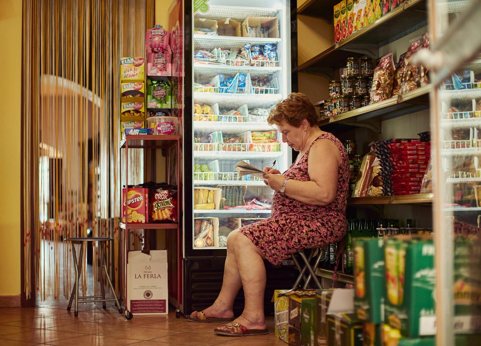 A woman in a pink floral dress sits in front of a refrigerated display case in a grocery store, surrounded by various food and beverage products.