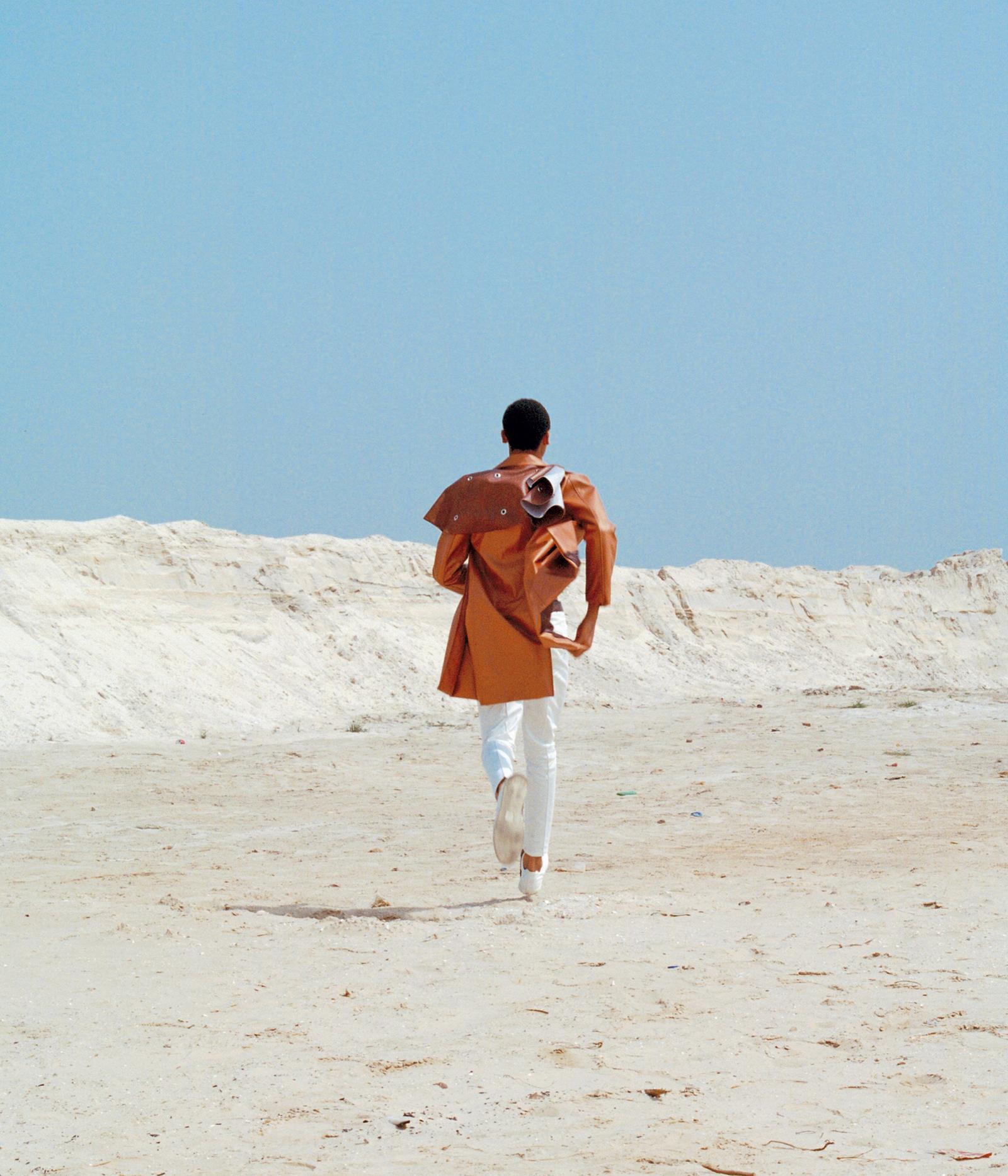 A man in a long, orange, leather jacket and white pants walking away from the camera in a desert-like environment.