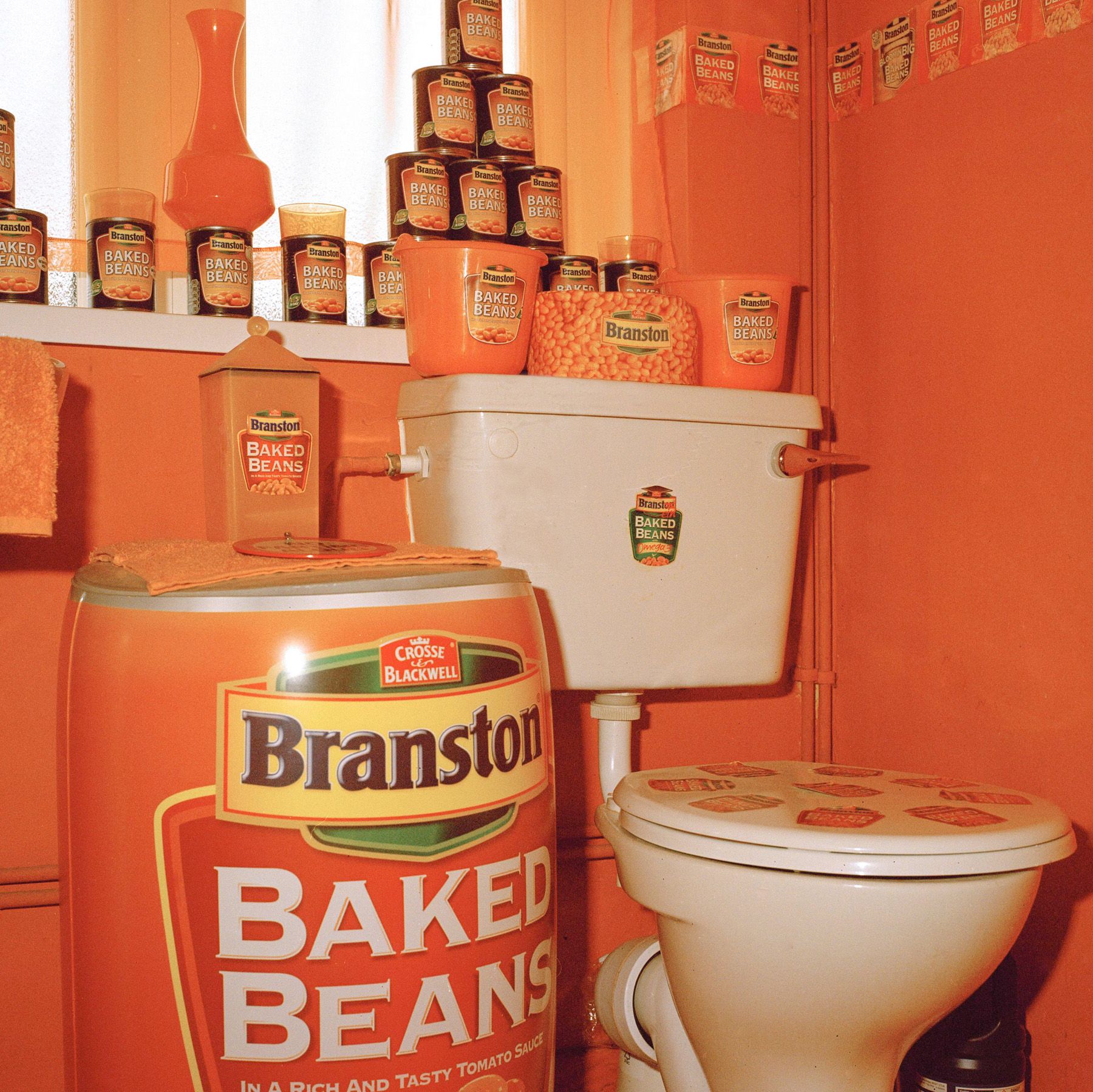A bathroom with large and small cans of baked beans arranged on shelves and near the toilet.