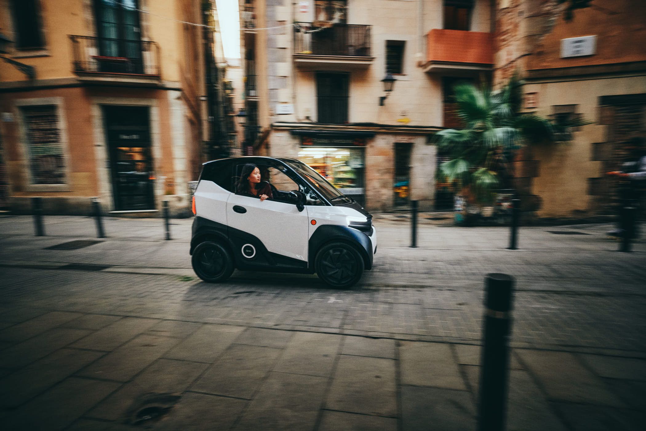 A small white car drives through a narrow, historic European city street, with a woman behind the wheel and a man in the passenger seat. The mood is lively, with a sense of urban.