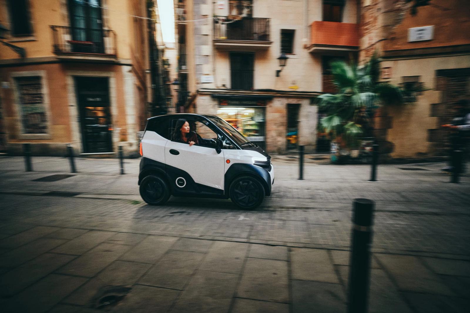 A small white car drives through a narrow, historic European city street, with a woman behind the wheel and a man in the passenger seat. The mood is lively, with a sense of urban.