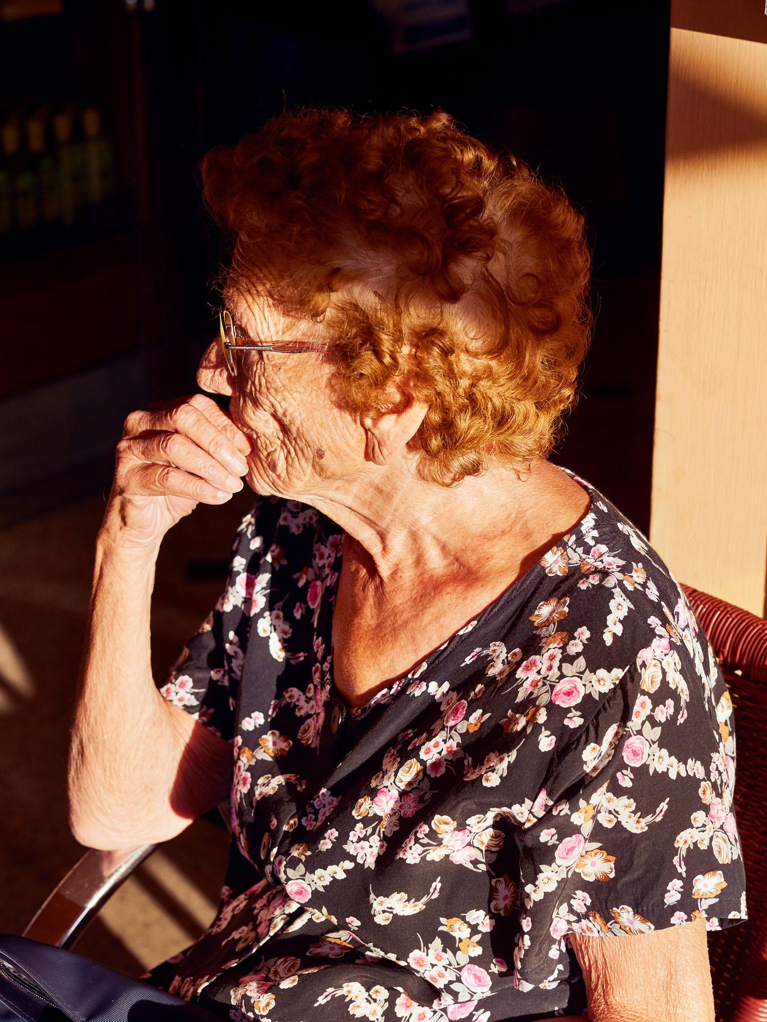 An elderly woman with curly red hair and a black floral blouse sits in a chair, looking out the window.