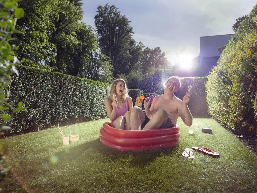 A man and a woman are sitting in a red inflatable pool in a backyard, surrounded by trees and a hedge, both holding ice cream cones and laughing.