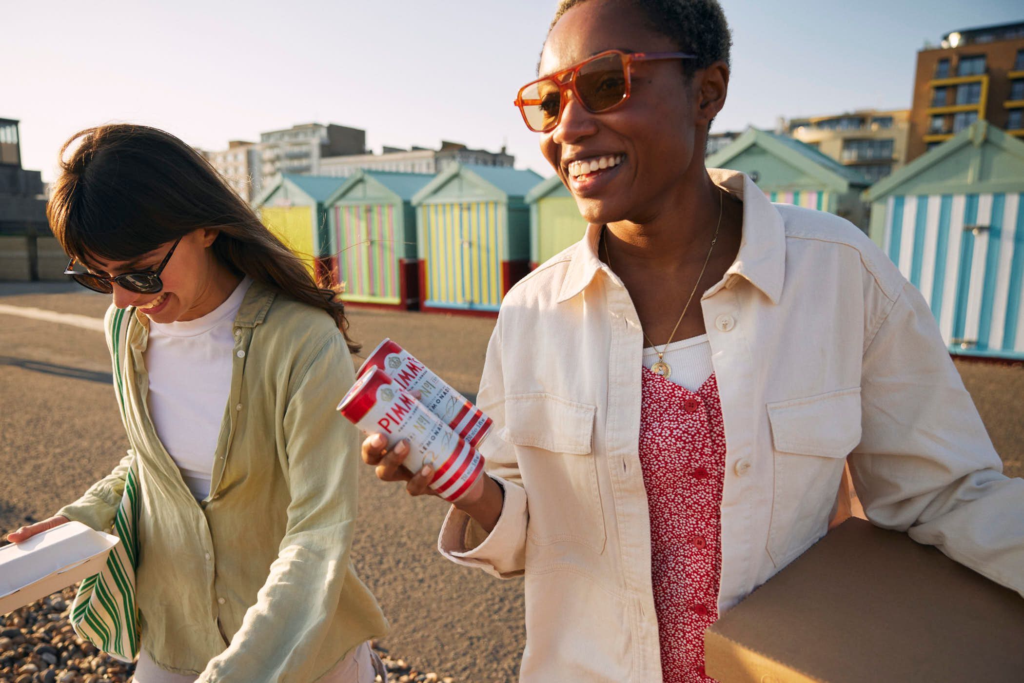 Two women are walking along a beachfront, carrying food and drinks in takeout containers. The woman on the right is wearing a white jacket over a red and white patterned top,.