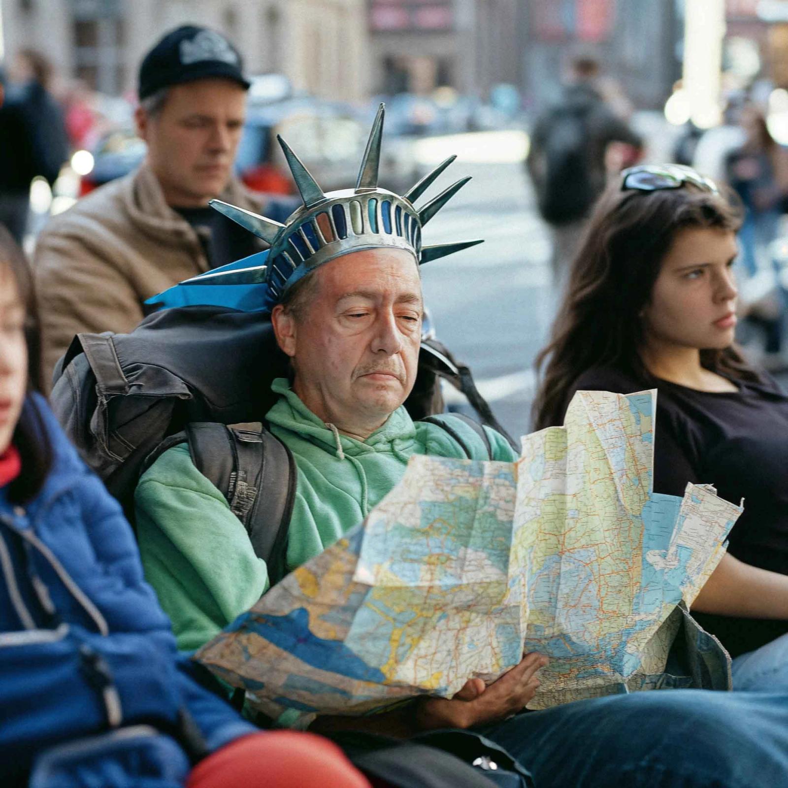 A man in a Statue of Liberty costume, wearing a green hoodie and backpack, holds a map and looks around in a crowded city street.
