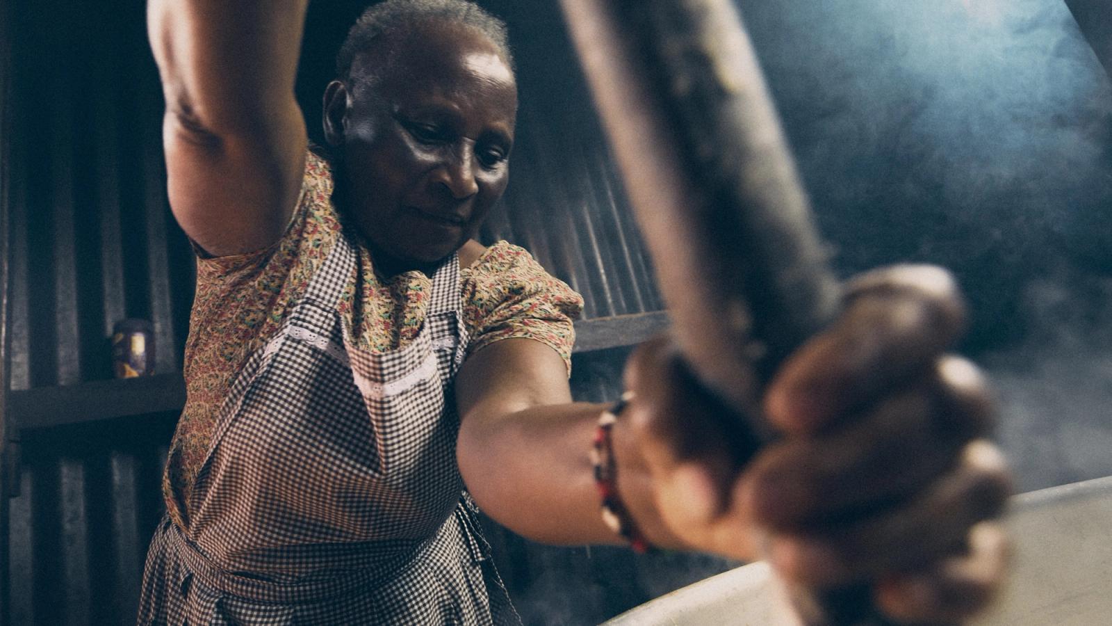 A woman in a floral blouse and a black and white checkered apron is stirring a large pot with a wooden spoon.