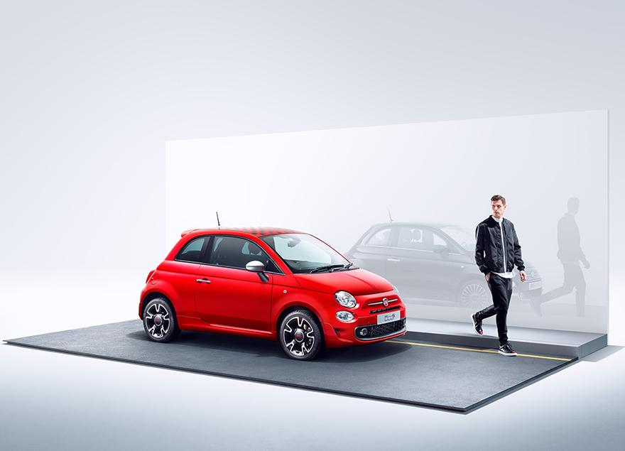A red Fiat 500 car with a man in a black jacket and black pants standing next to it, on a grey background.