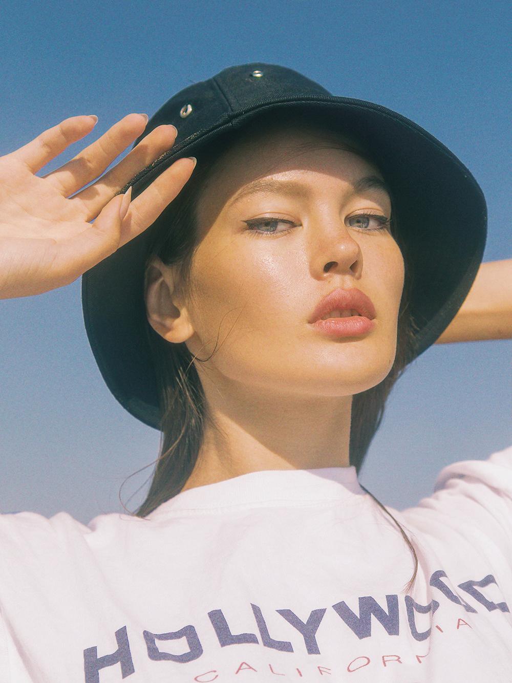 A woman wearing a black hat and a white t-shirt with the word 'Hollywood' on it, posing in front of a blue sky.