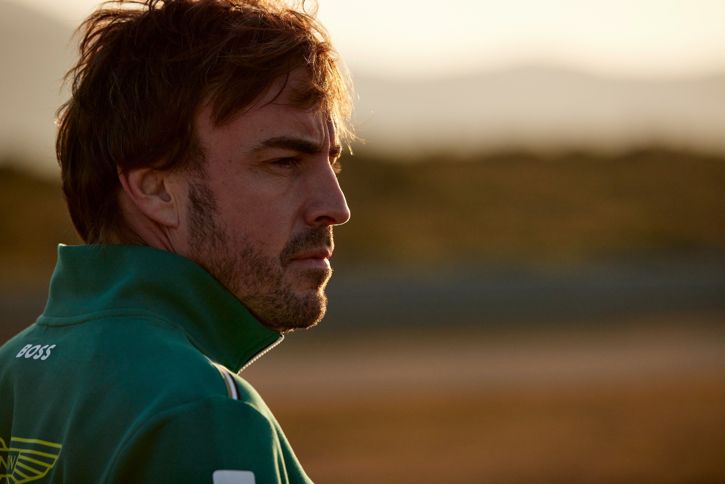 A man with a rugged, weathered face and a hint of a smile, wearing a green Aston Martin racing team jacket, with a blurred background of a racing track or a similar environment.