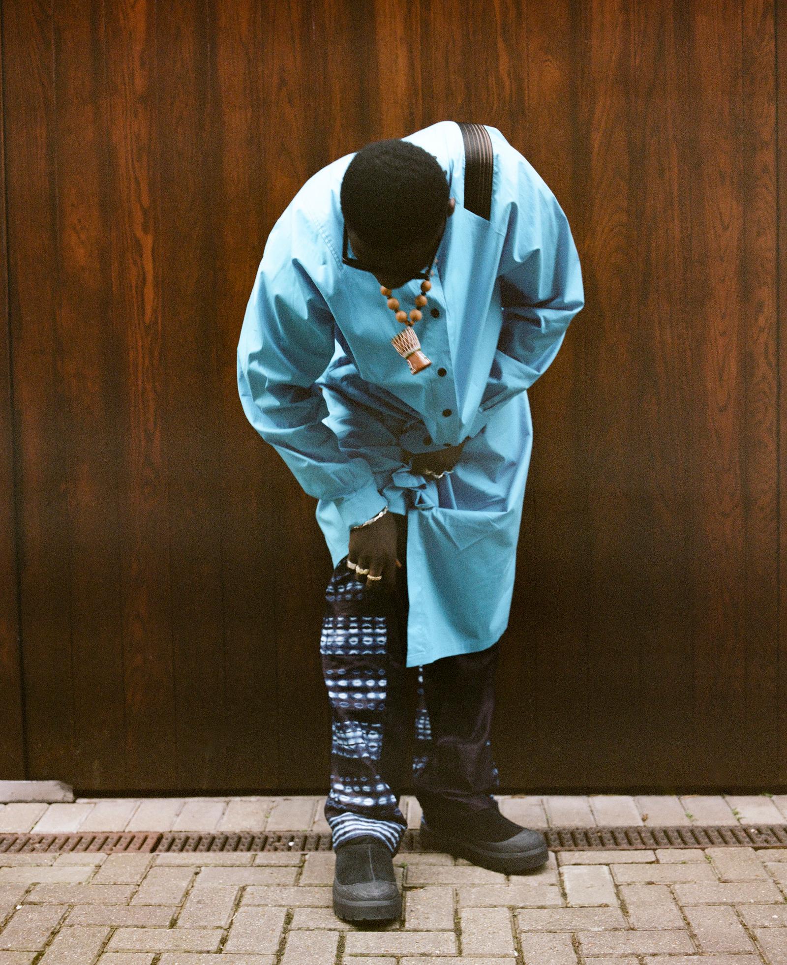 A man in a blue trench coat and patterned pants is standing in front of a dark wood panelled wall, looking down at his shoes.