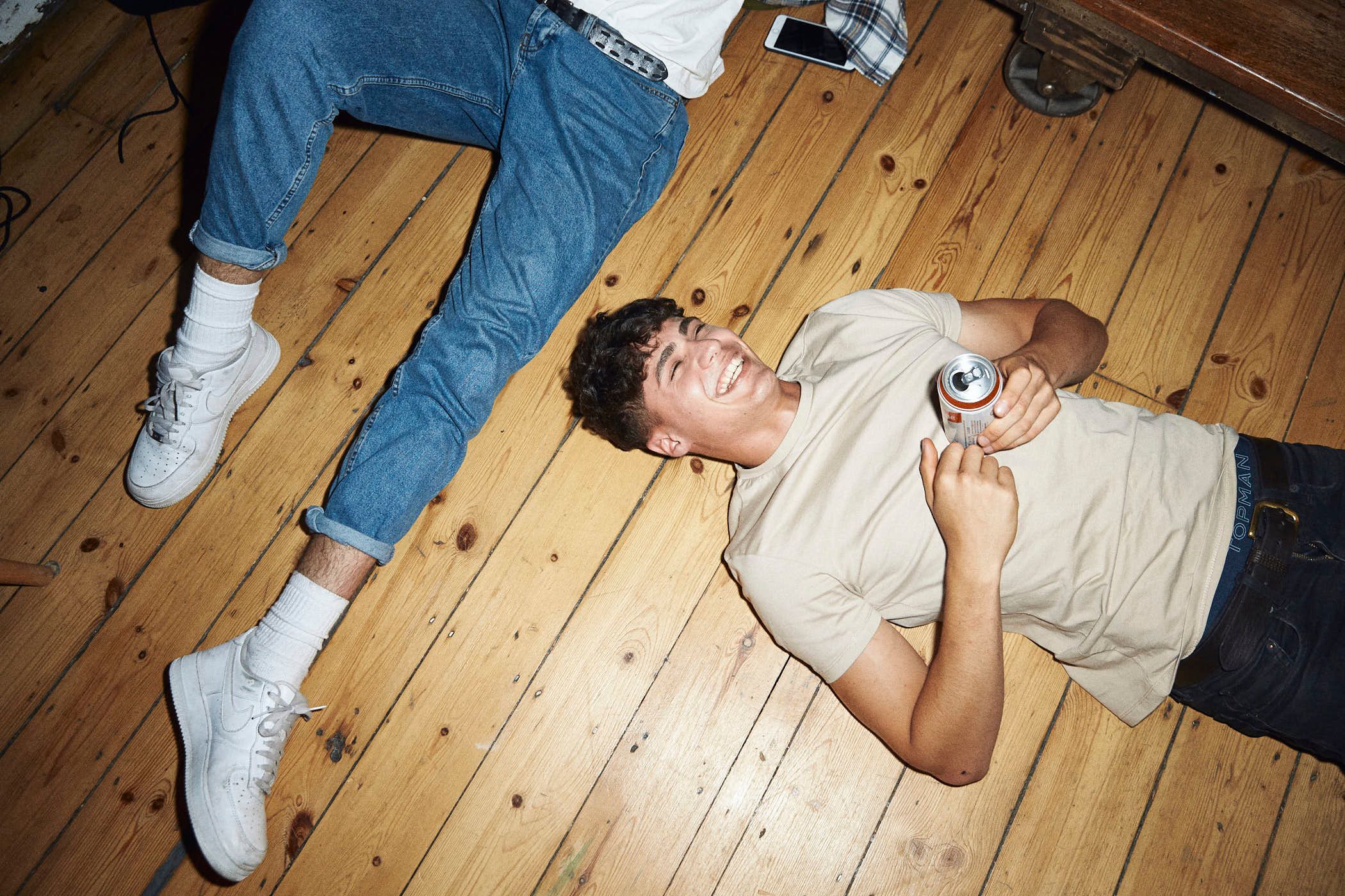Two young men lying on the floor, laughing and holding a can of beer. They are both wearing casual, comfortable clothing and appear to be enjoying themselves.