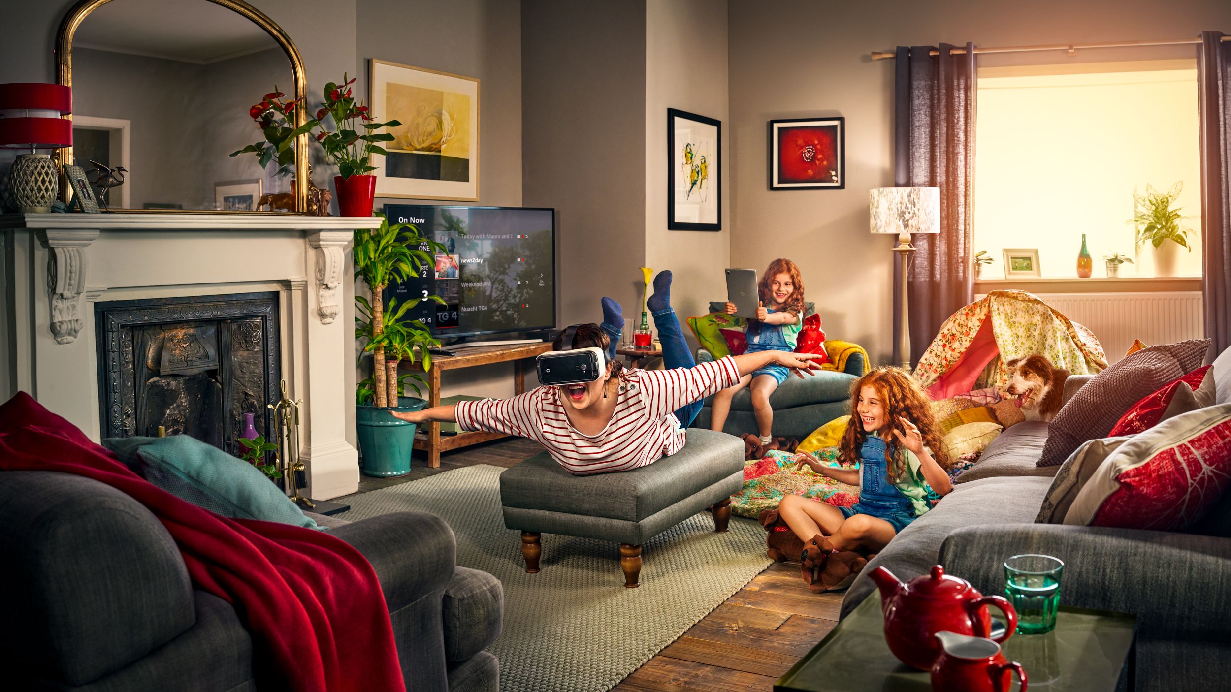 A woman wearing a VR headset and a man sitting on a couch, both surrounded by a living room with a TV, a fireplace, and a few pieces of furniture.