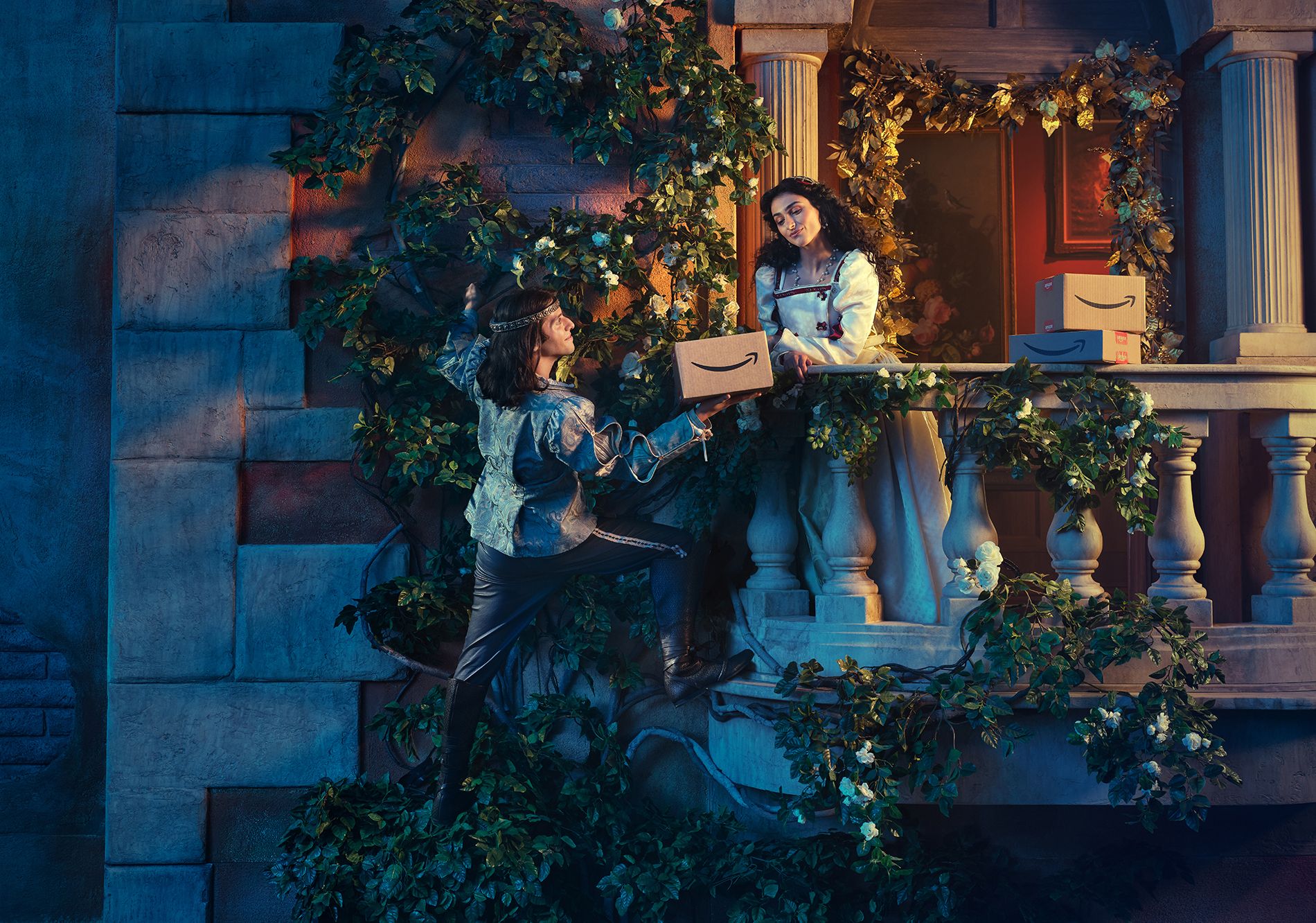 Two women in Renaissance-style clothing, one holding a box with the Amazon logo, standing on a balcony with a stone wall and columns, surrounded by greenery and flowers.