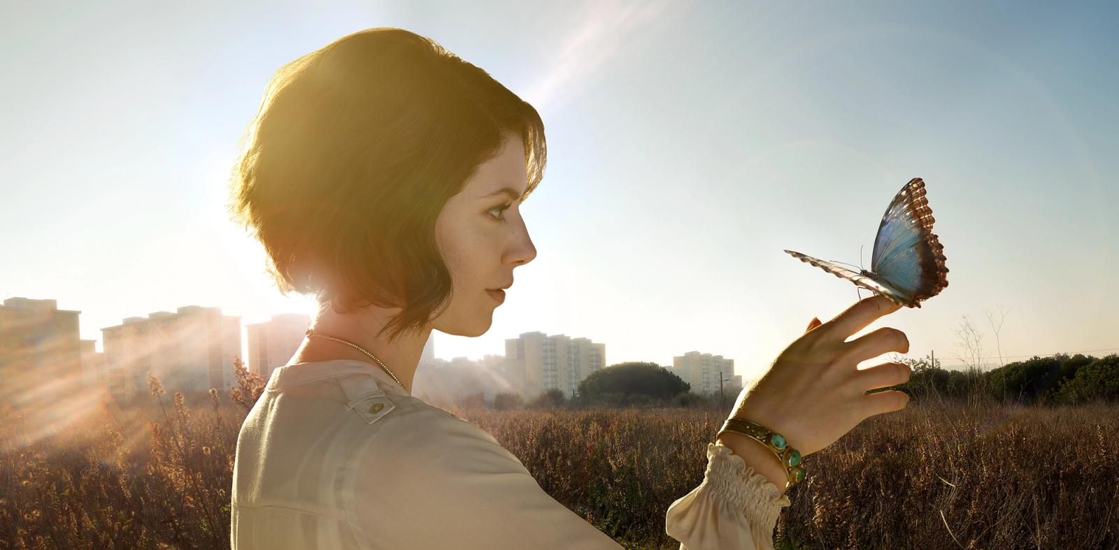 A woman with short, dark hair and a white shirt is standing in a field, with a butterfly perched on her hand.