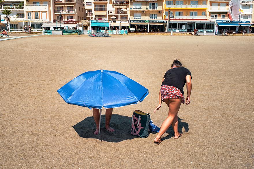 Two people are standing on a beach, one is holding a blue umbrella and the other is walking away from it.