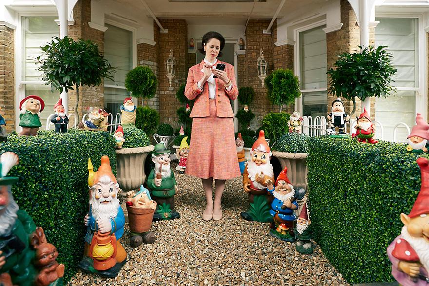 A woman in a pink tweed suit standing in front of a house with a garden full of garden gnomes.