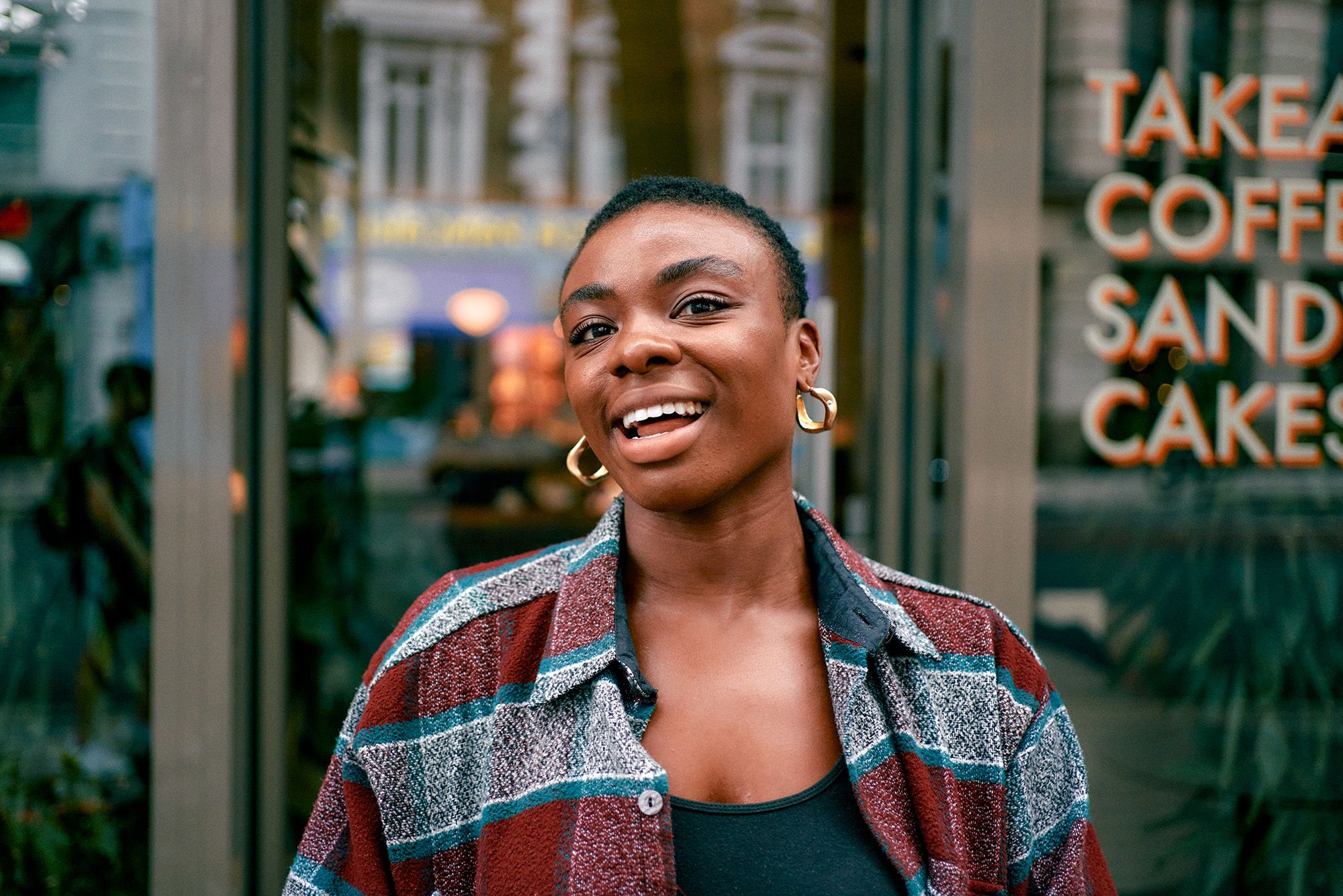 A woman with short hair and a warm smile, wearing a red and blue plaid shirt and a black tank top, standing in front of a city street with a blurred background.