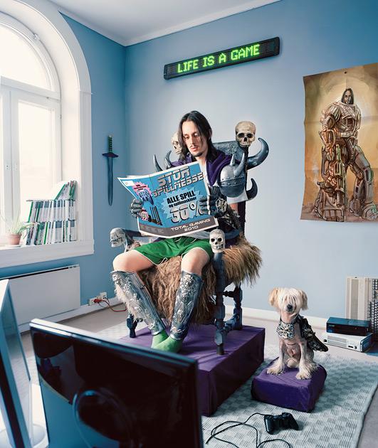 A man sits in a chair with a magazine, surrounded by objects and a dog.