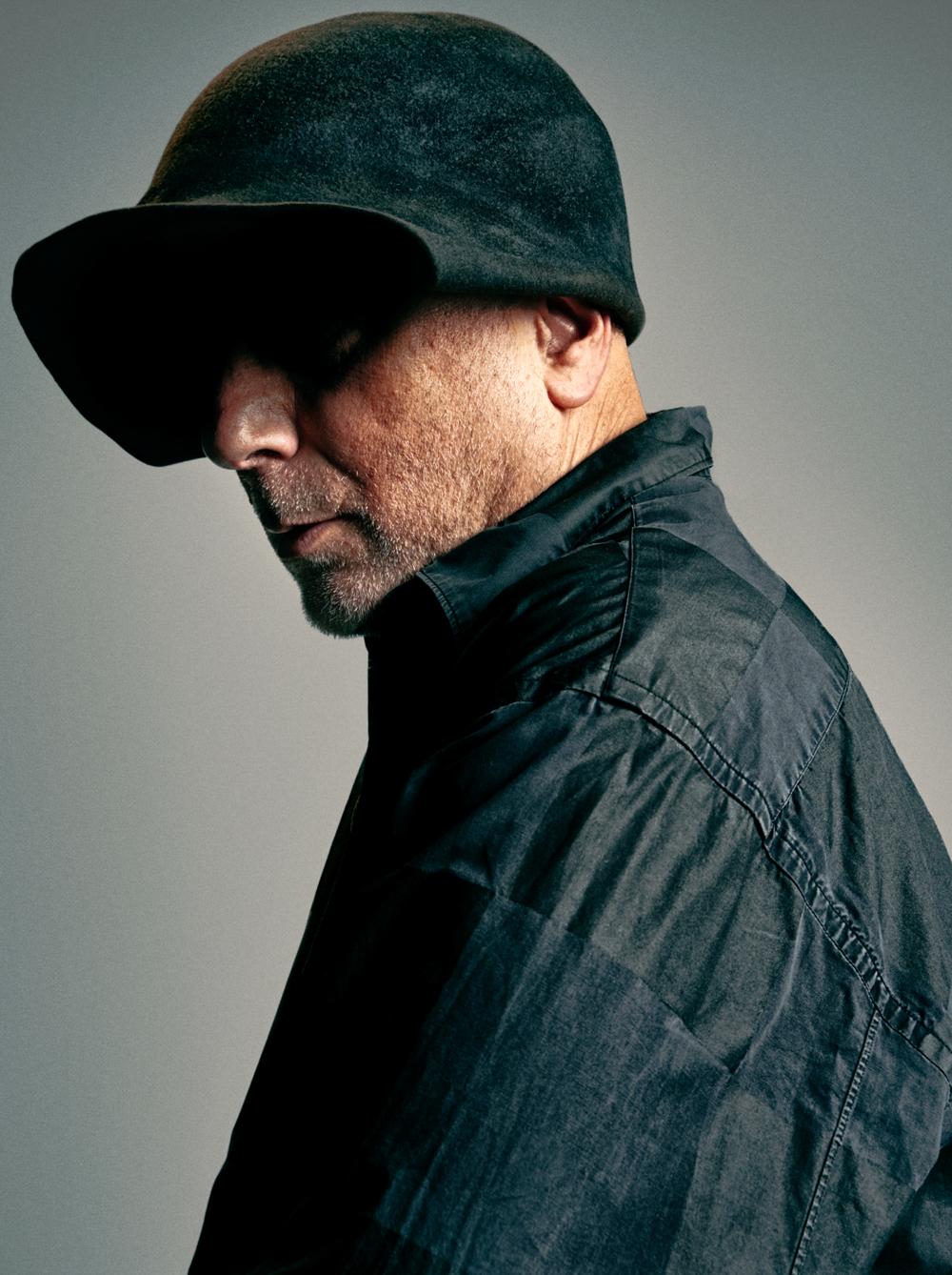 A man wearing a black fedora hat and a black leather jacket, looking over his shoulder with a serious expression.