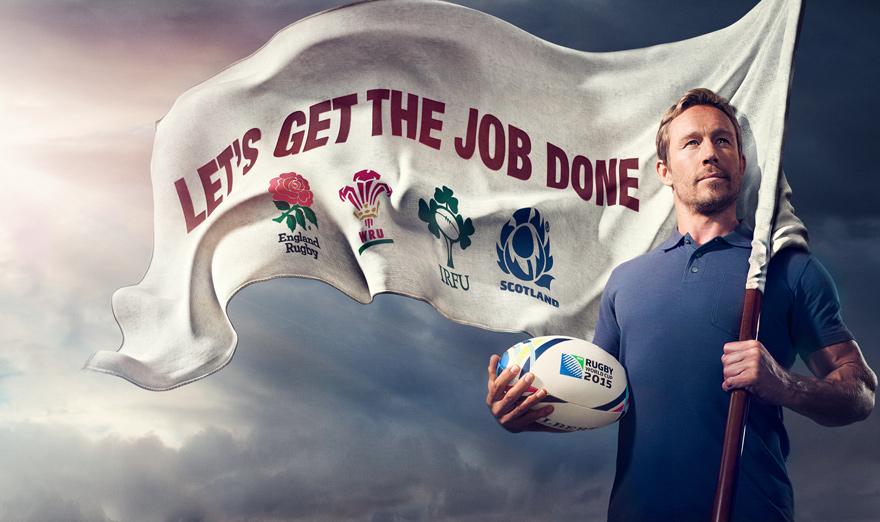 A man holding a flag with a football (or rugby) team's logo and a football (or rugby) ball, in front of a cloudy sky.