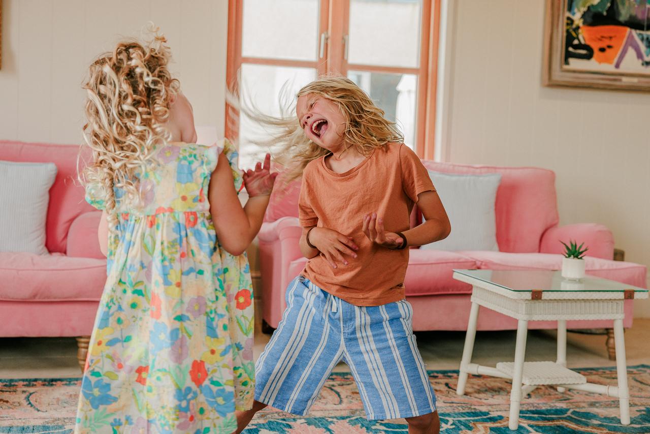 Two children dance in a living room with a pink sofa and a large window.