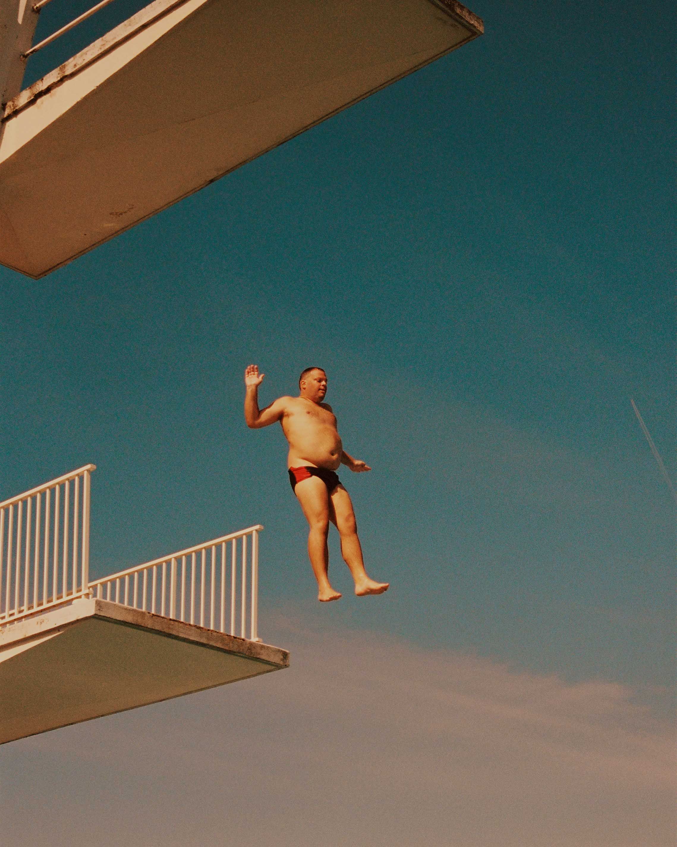 A man in a red and black swimsuit is in mid-air, performing a dive from a high platform into a pool.