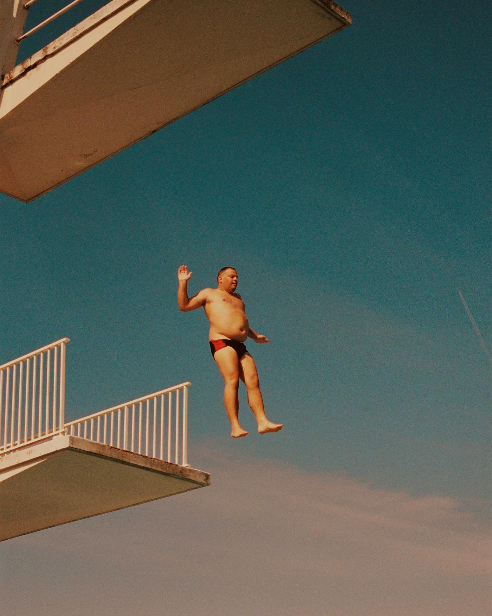 A man in a red and black swimsuit is in mid-air, performing a dive from a high platform into a pool.