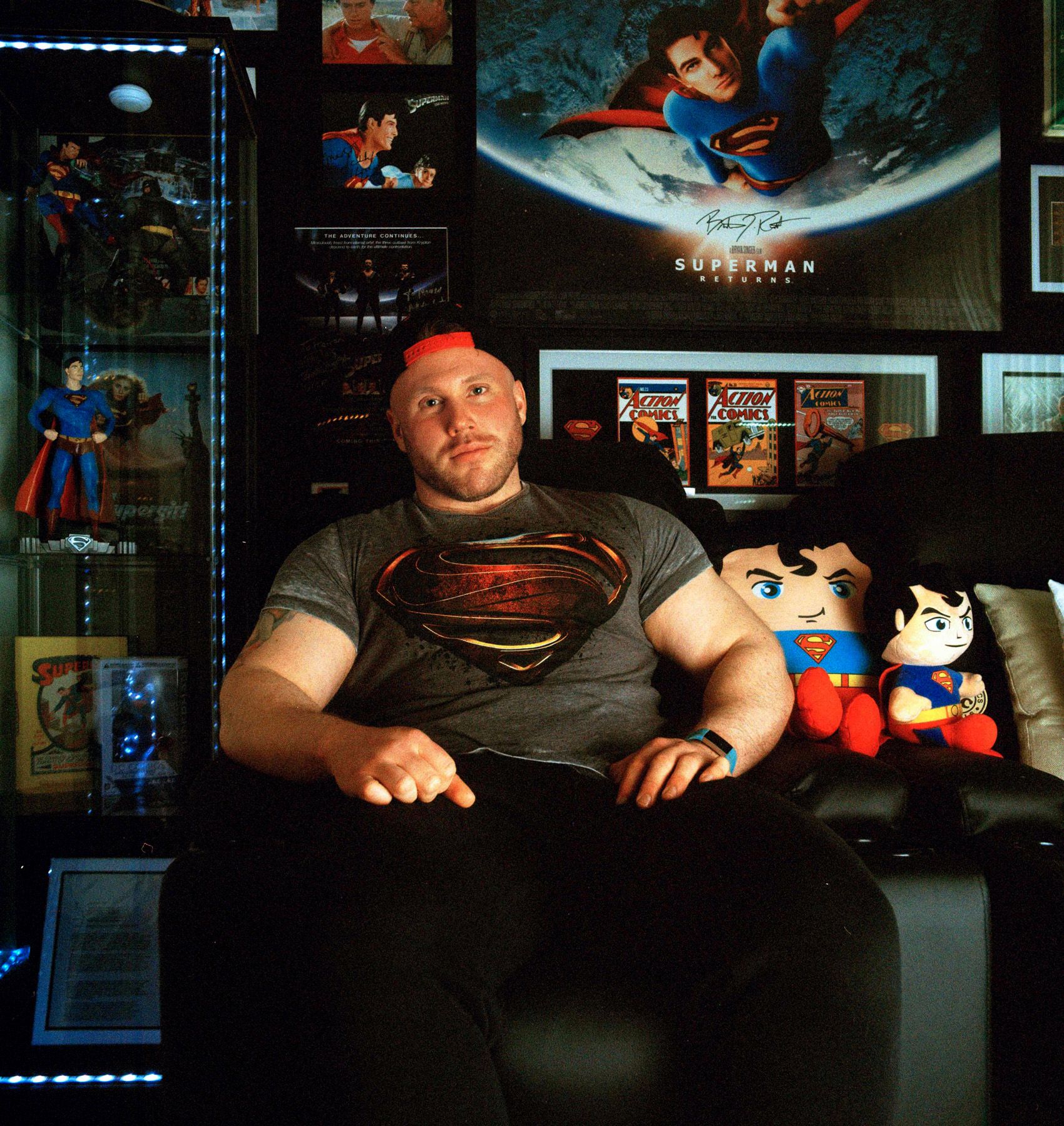 A man sits in a room with Superman memorabilia, wearing a Superman t-shirt and jeans.