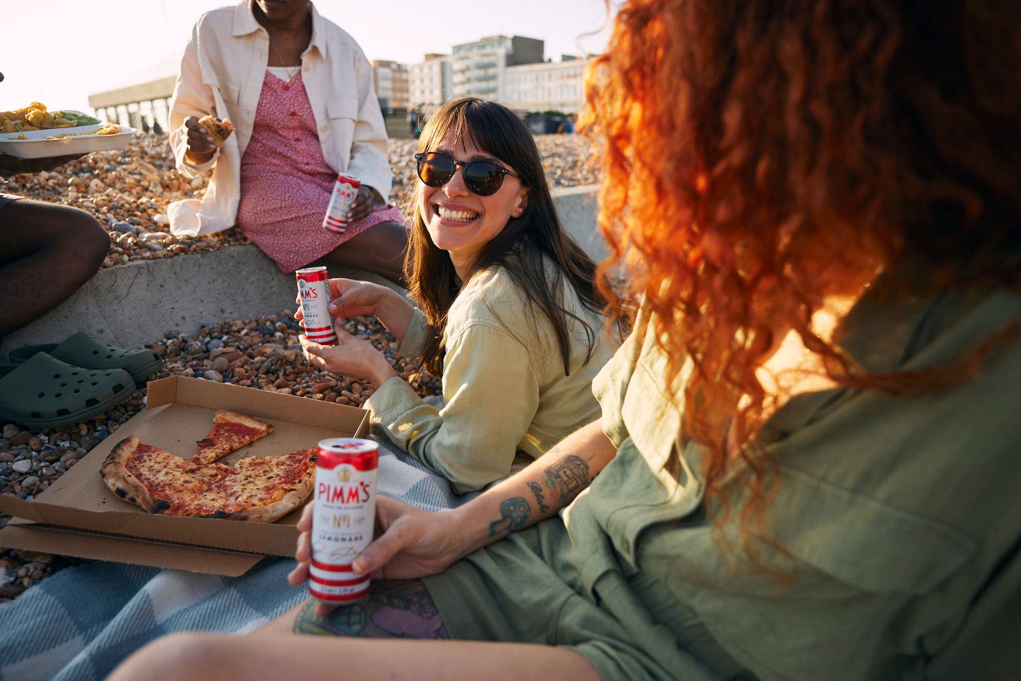 A group of women sit on a beach, enjoying pizza and Pimm's, a popular British summer drink. The woman in the foreground, with long red hair, holds a Pimm's can, while others in.