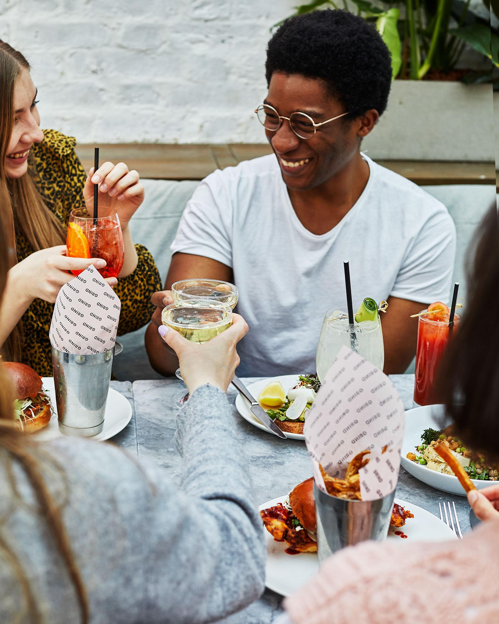 A group of people, including a man with a white t-shirt, are enjoying food and drinks at a table, with multiple plates of food and several drinks in front of them.