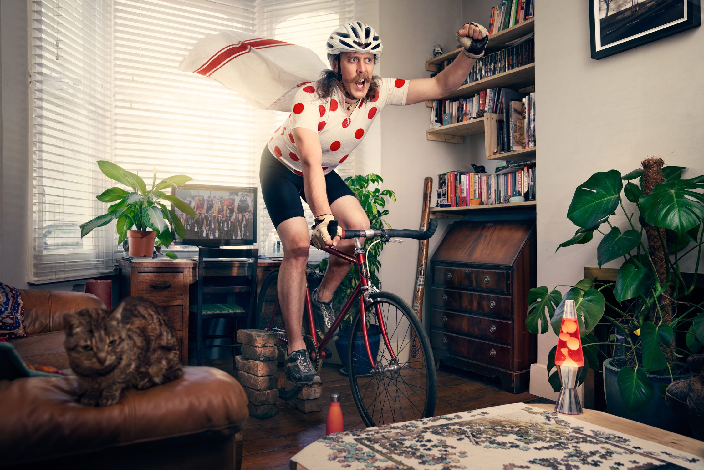 A man rides a red bicycle in a living room with cats.